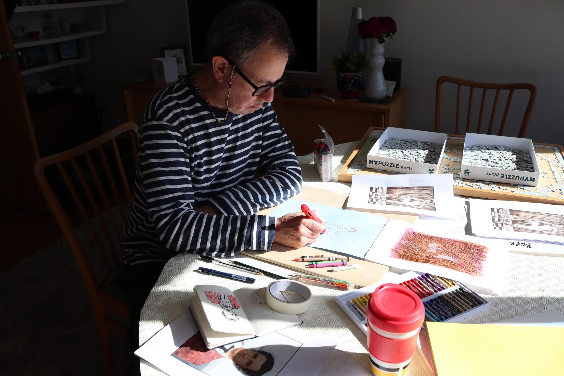 A man in a blue and white striped long sleeved tshirt sits drawing at a table covered in notebooks and coloured pencils