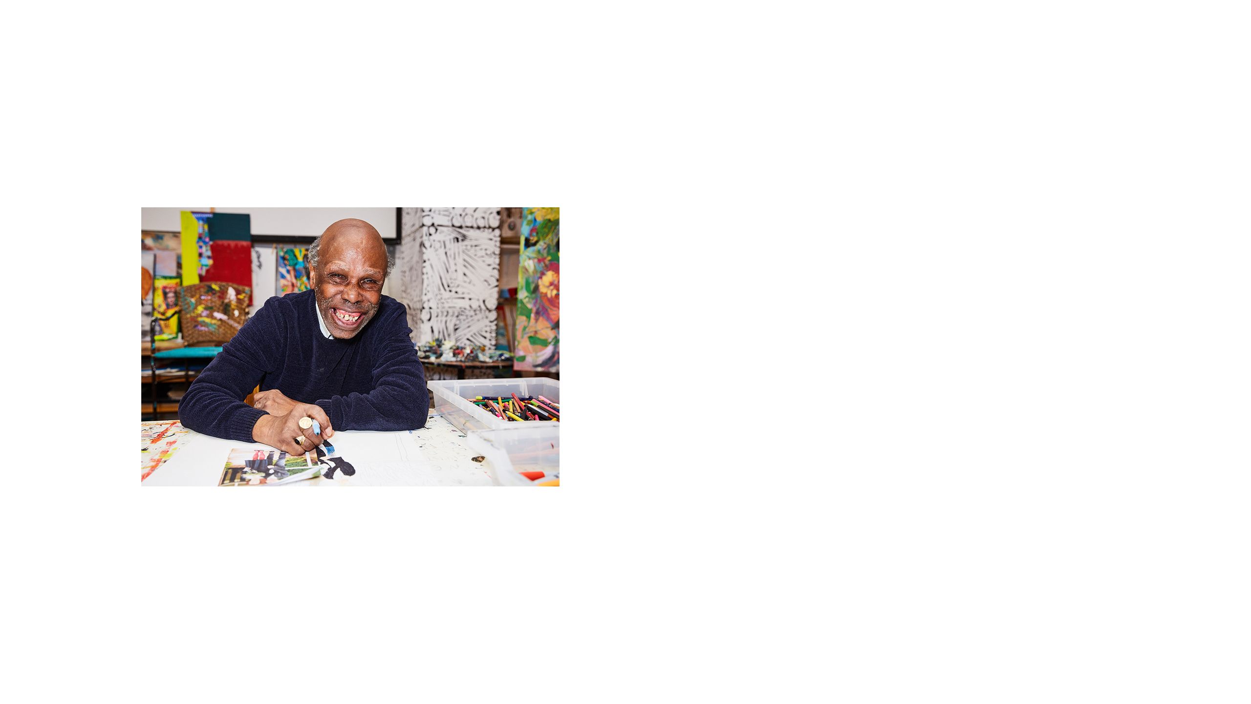 A man with a big smile sits at a desk holding a pen, drawing a colourful portrait