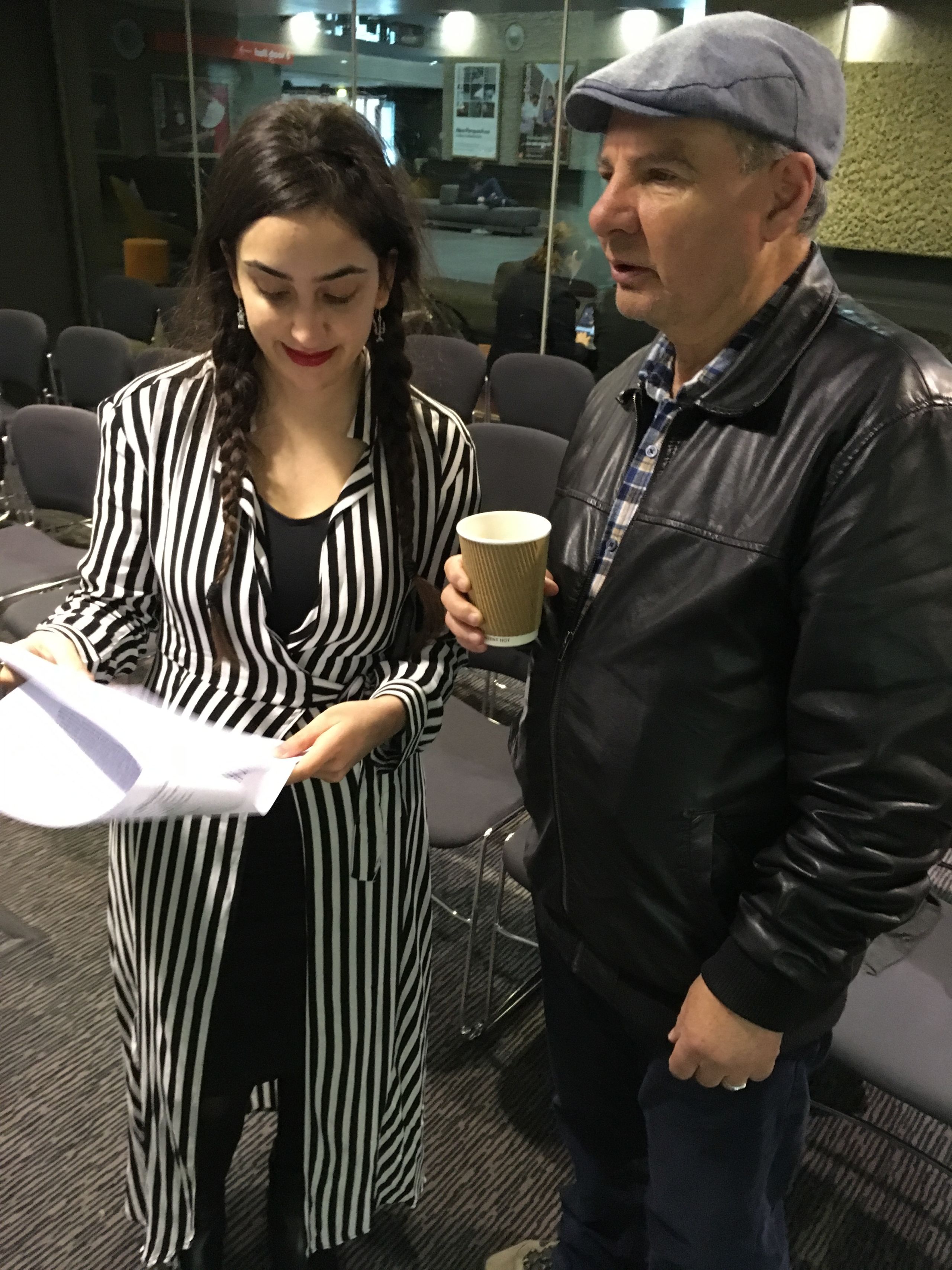A man in a flatcap holding a paper coffee cup stands next to a woman, wearing a black and white striped dress, who is holding a piece of paper