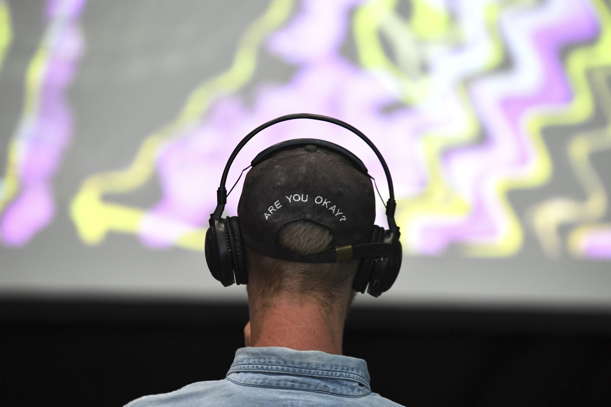 The back of a man's head, sitting in front of a large television screen, he is wearing headphones and a baseball cap which has text saying 'Are you okay?'