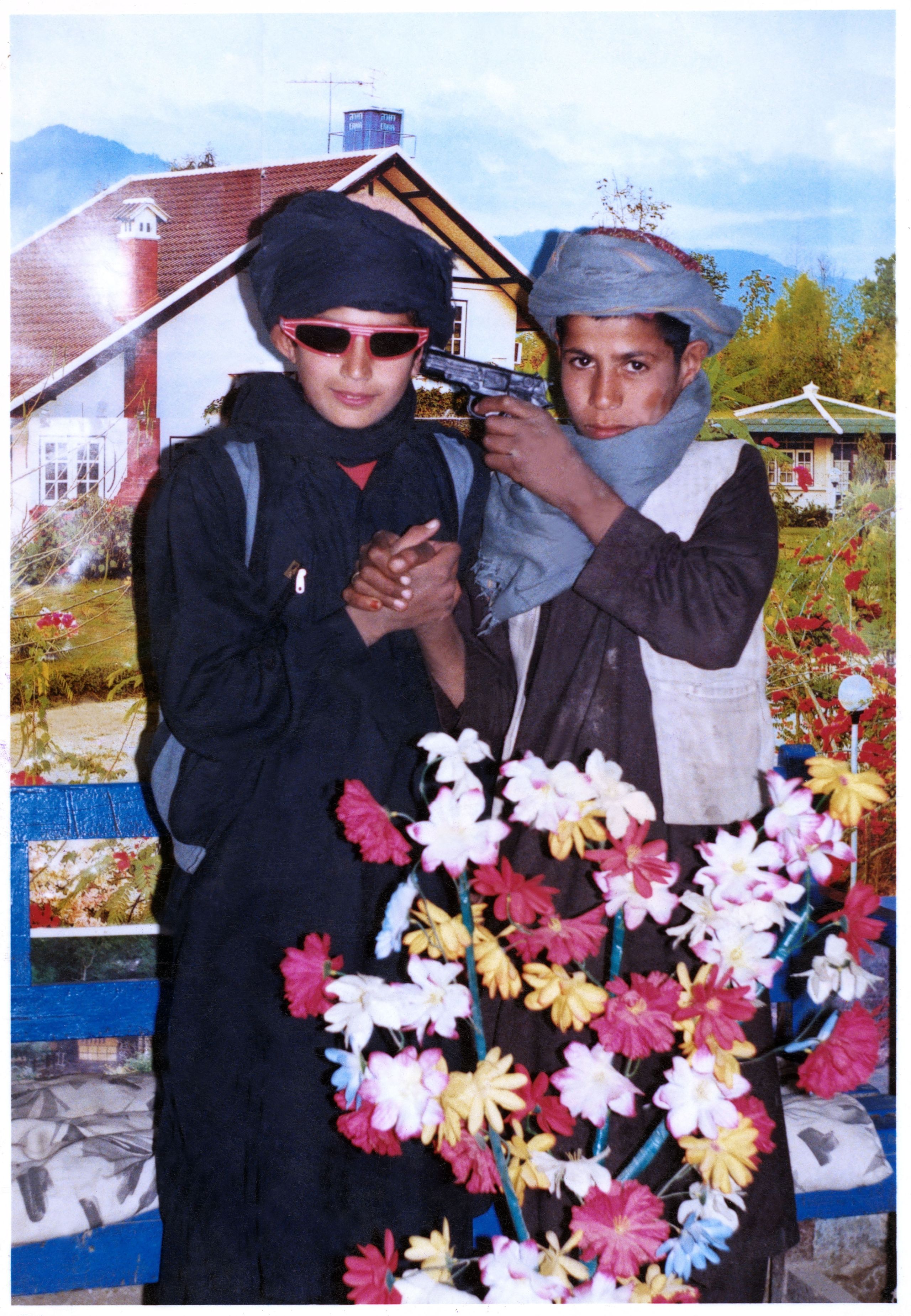 Two Afghani Boys hold hands, posing before a photographer's Swiss chalet backdrop. One boy holds a gun to the other boy's temple