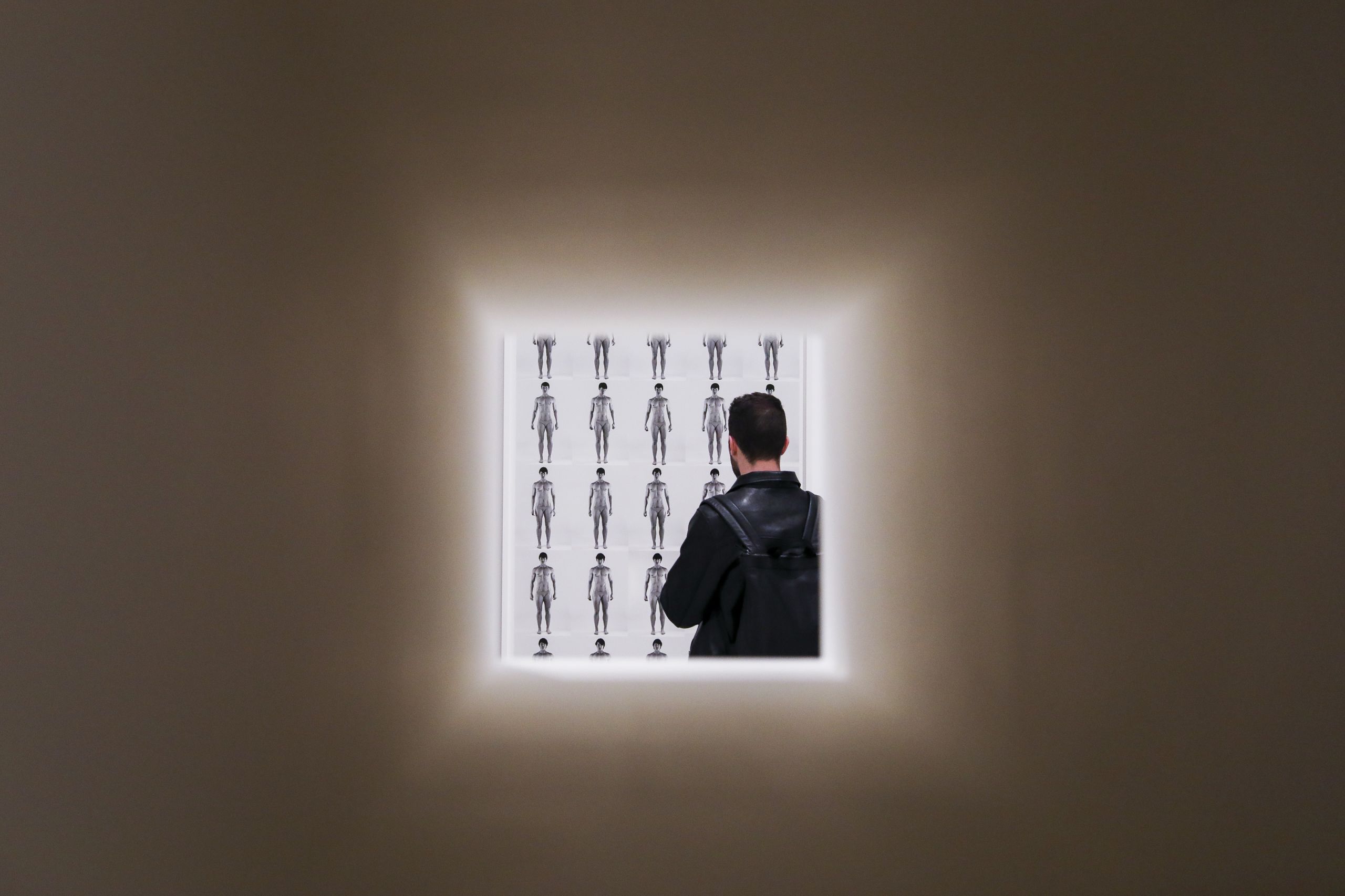 A man in a black jacket with a backpack looks at a black and white image featuring a grid of timelapse photographs of a muscular figure. The image is framed in a square, with the photograph taken through a hole in the gallery wall.