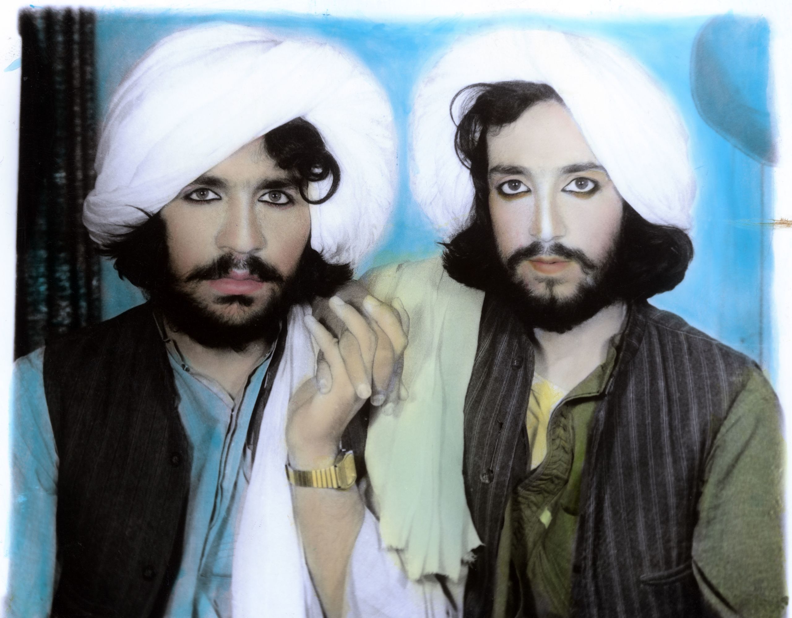 A colour photograph of two Afghani men clasping hands. They wear white turbans, and their eyes are outlined with kohl.