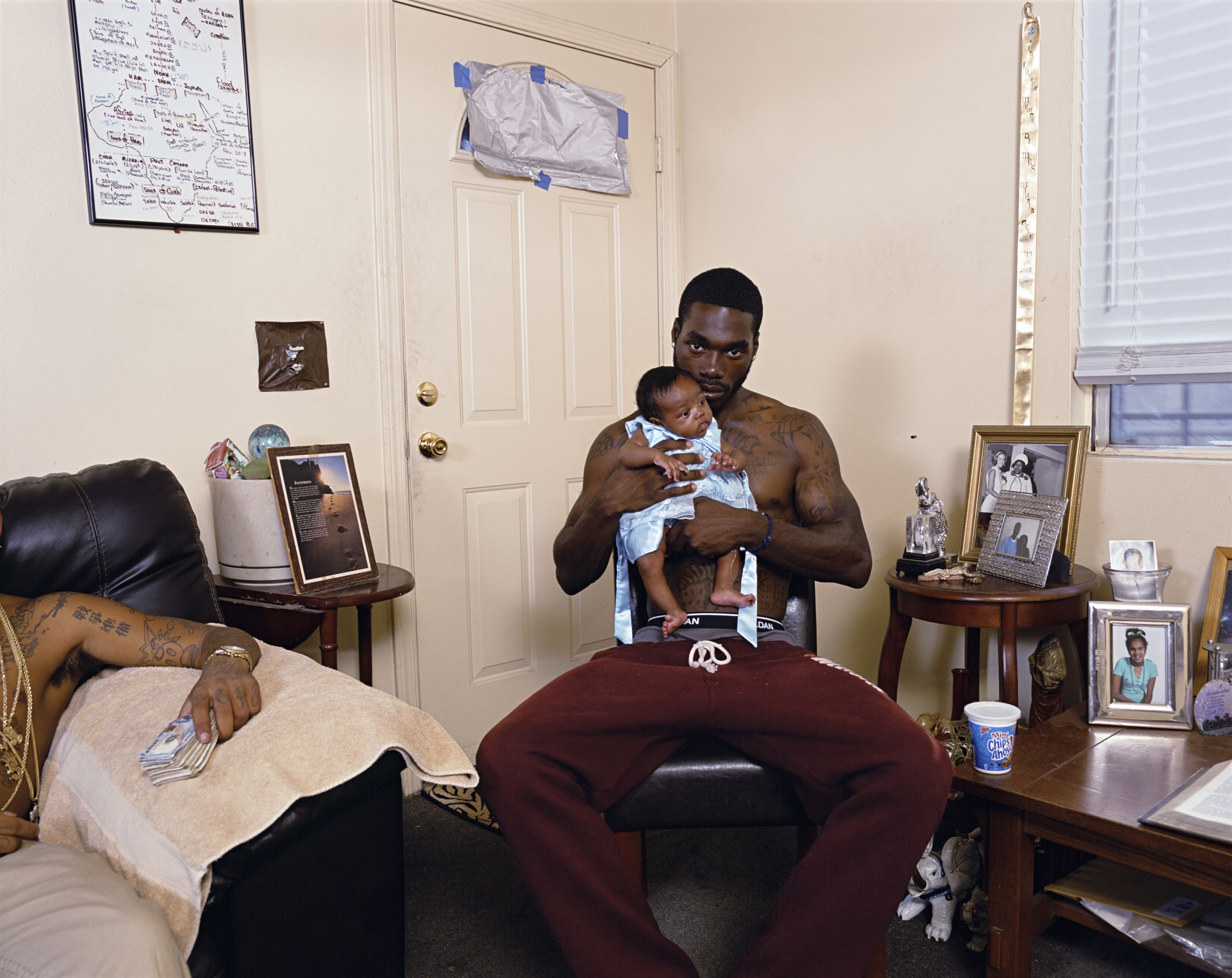 Large scale, colour photo graph of two black men in a domestic setting. the man at the centre of the photo holds a baby girl, while the second man, who is only partially visible holds a wad of cash in his outstretched hand.