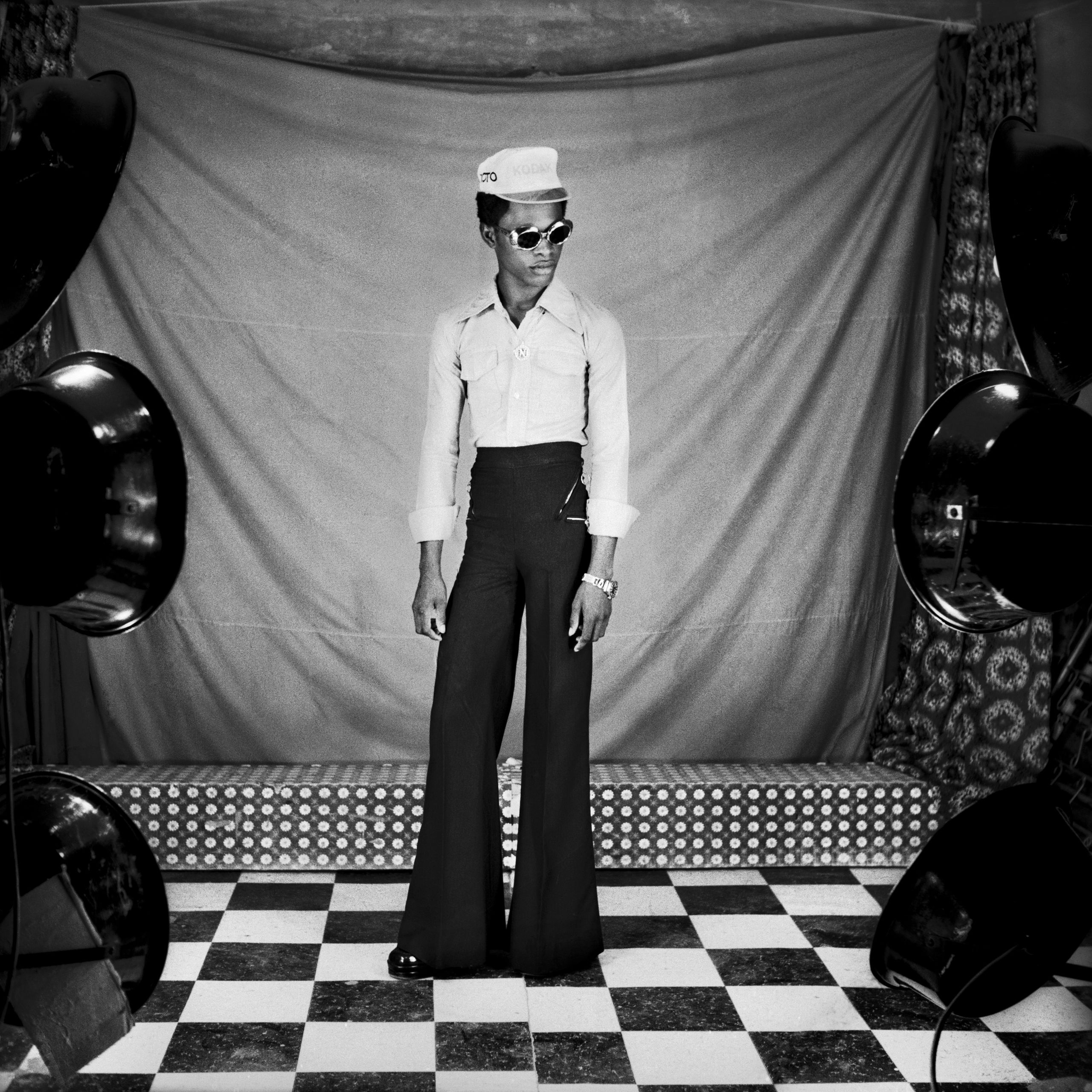 A man in a white shirt and black flares, wearing sunglasses and a white hat, stands on a stage with a checkerboard floor