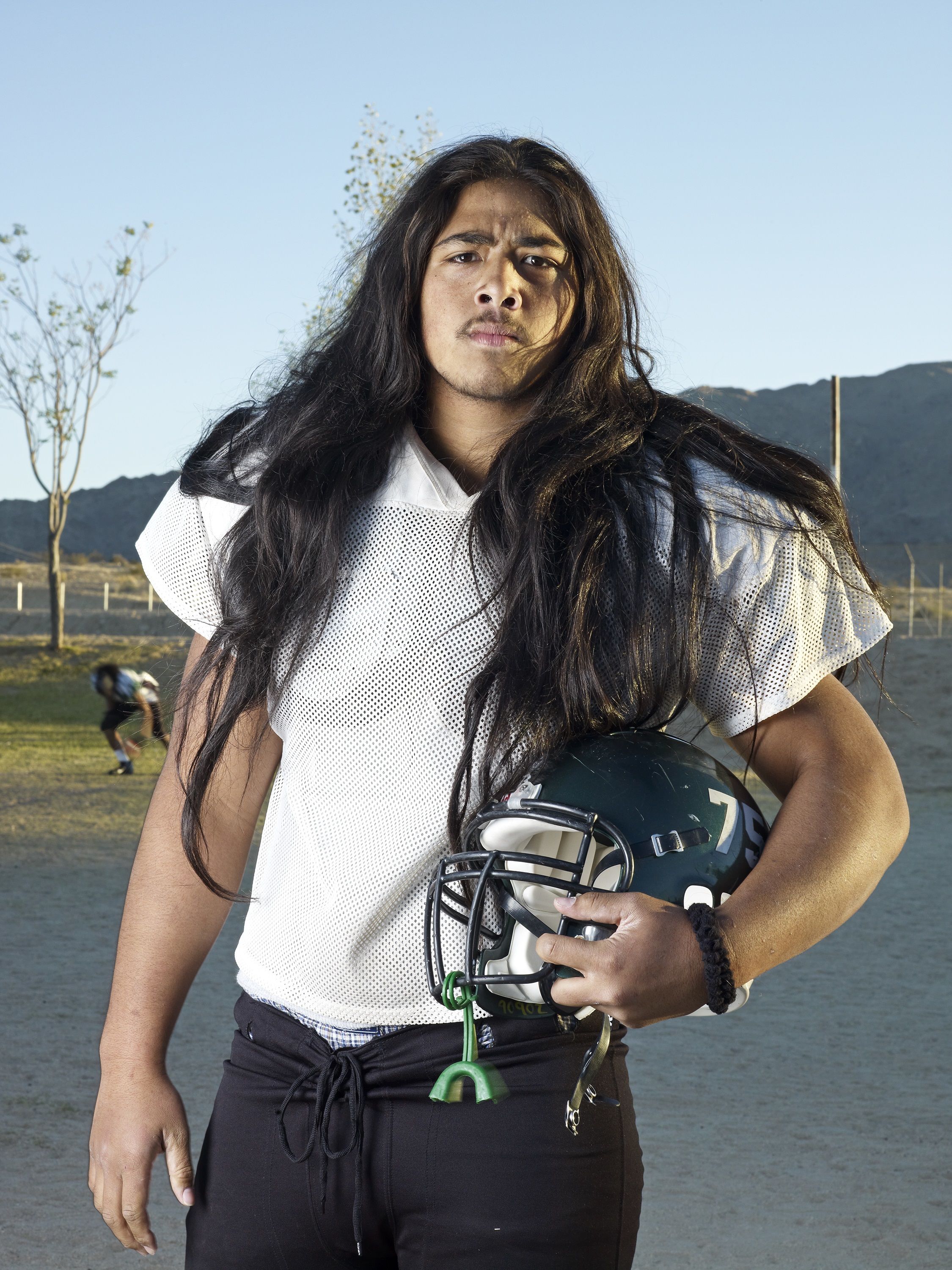 A man with long hair wearing a white t-shirt and holding an American football under one arm.