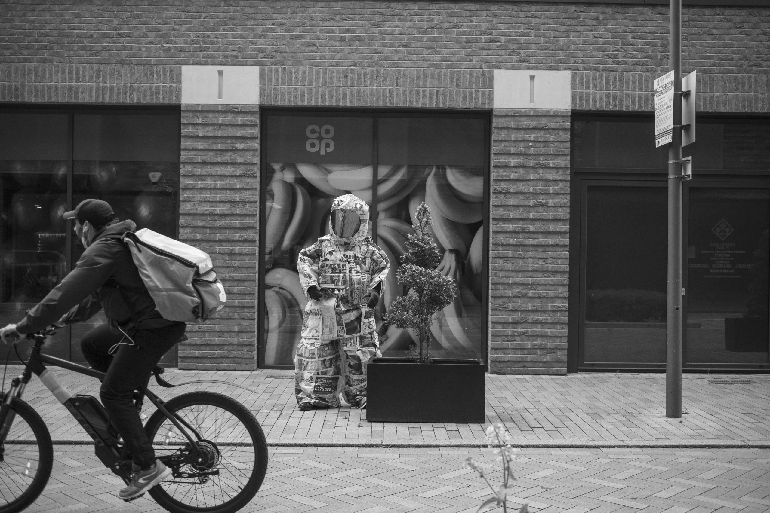 a black and white photo of a person wearing a spacesuit made of newspapers standing in front of the façade of a co-op supermarket, in the front a masked cyclist cycles past