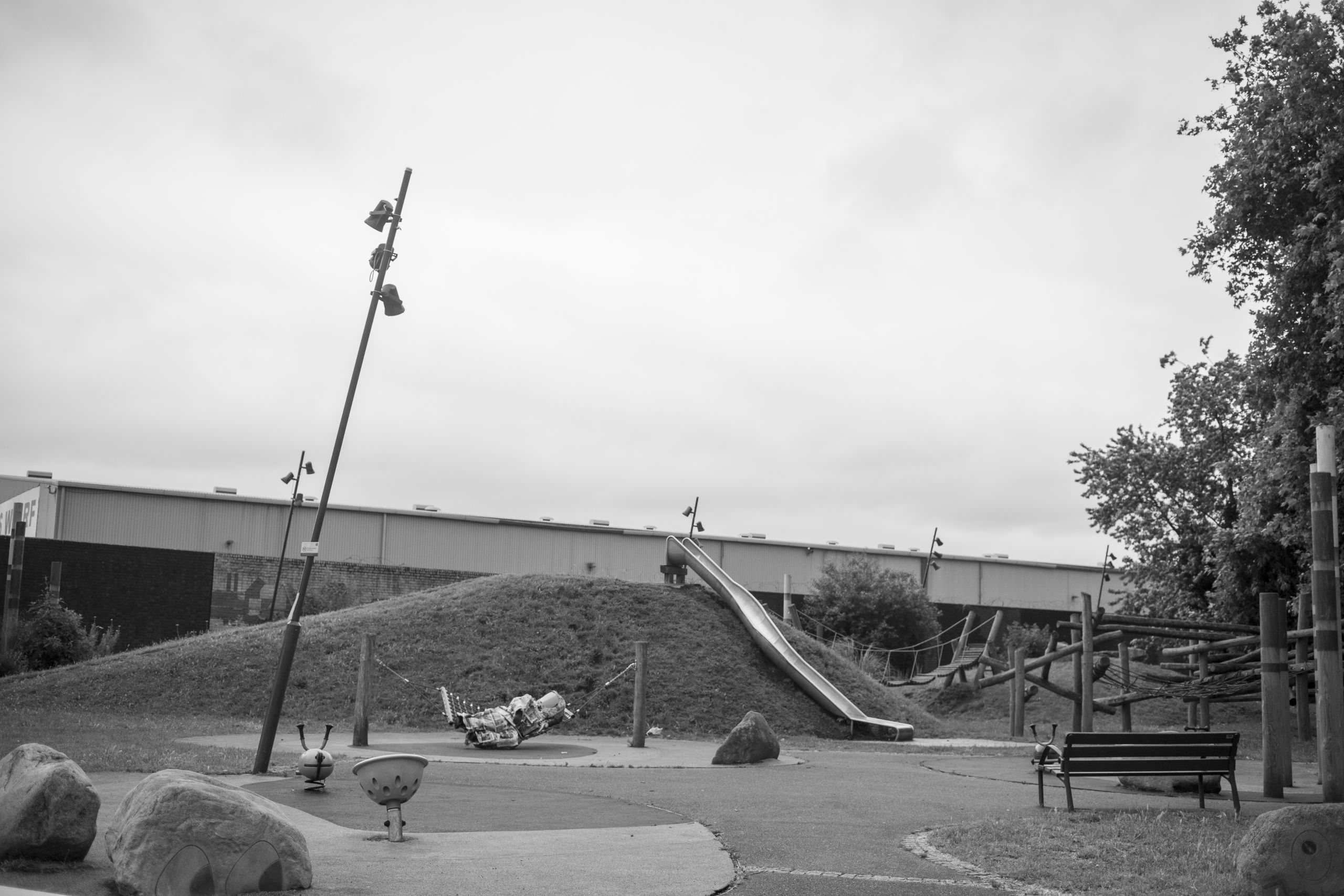 a black and white photo of a playground with a lamppost, a bench, and a slide on top of a little grass hill, someone wearing a spacesuit made of newspapers is lying on top of one of the play structures