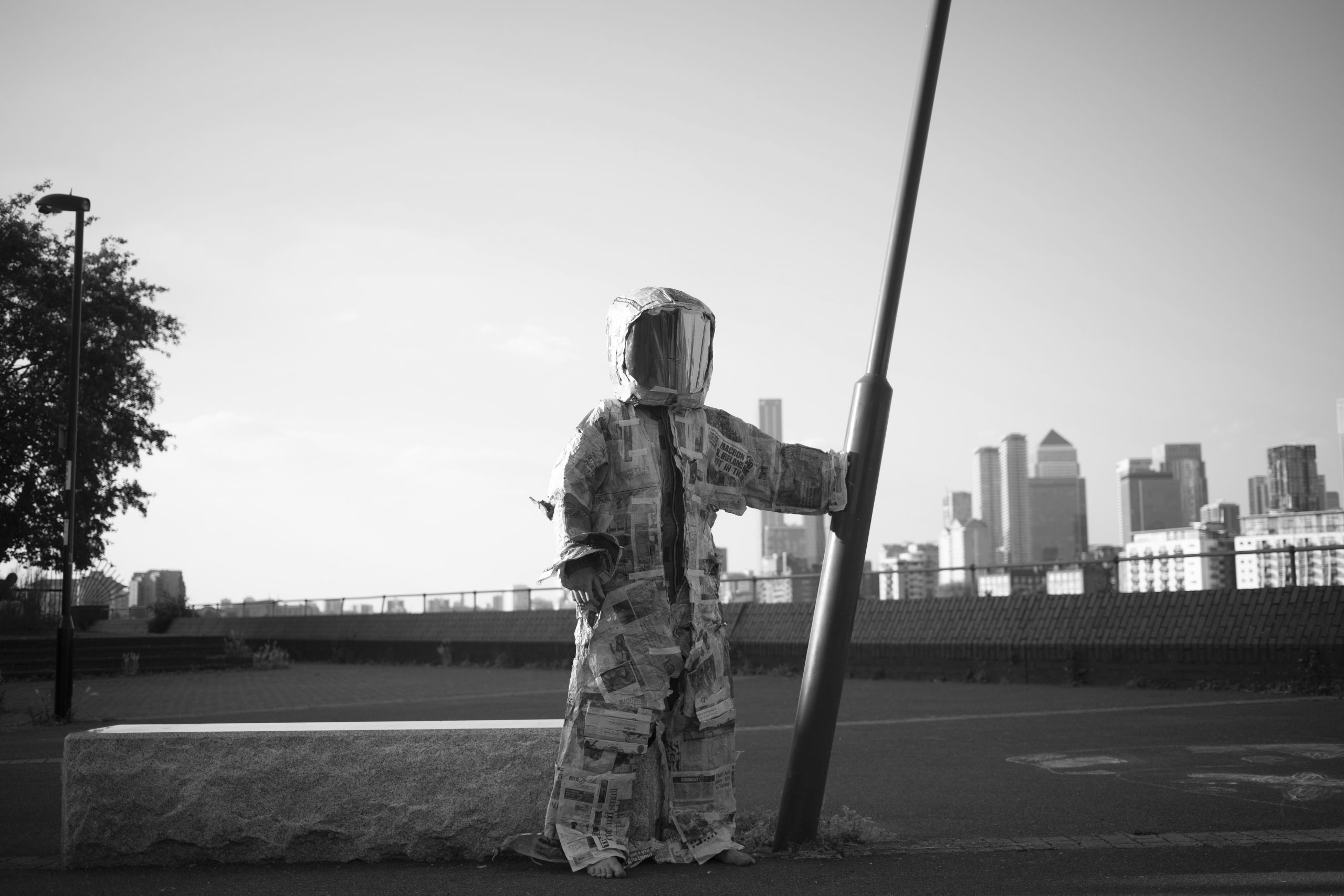 a black and white photo of a person wearing a spacesuit made of newspaper, they are on a deserted street against the backdrop of the city, and they are holding on to a leaning lamppost