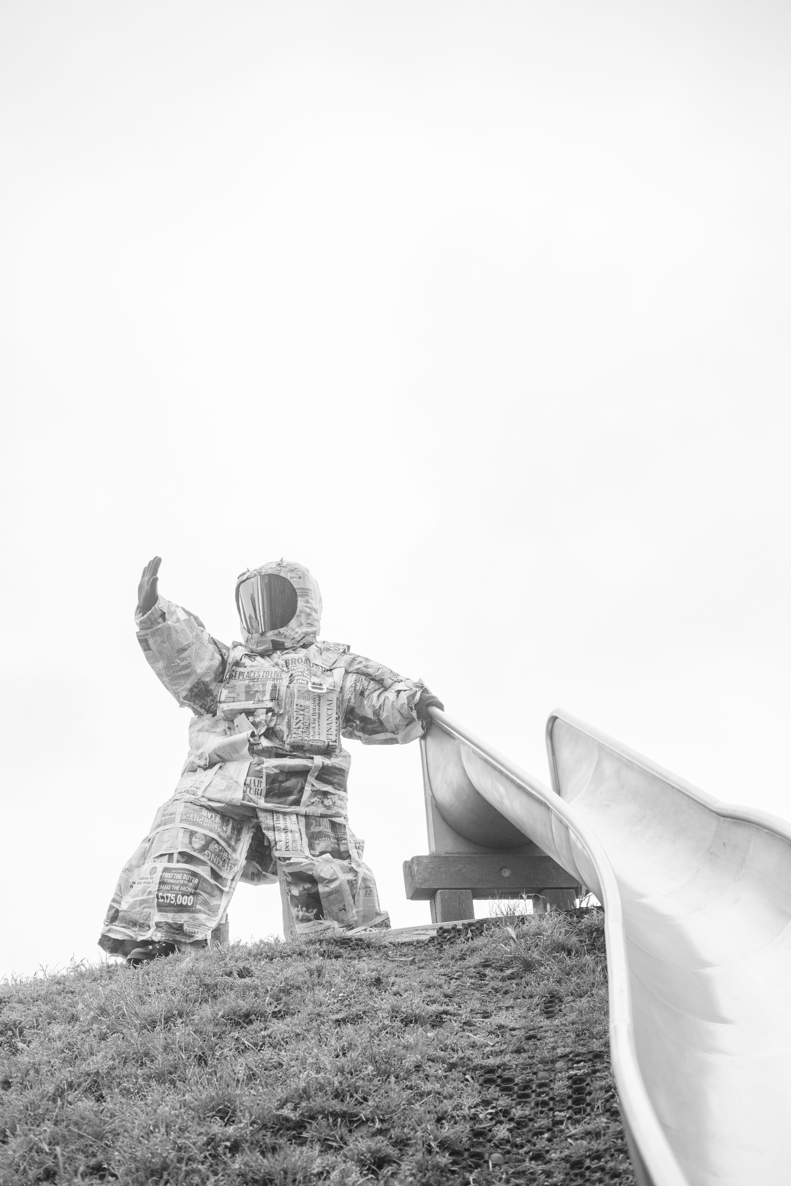 a black and white photo of someone wearing a spacesuit made of newspapers, standing on top of a grass hill, one hand on the top of a slide, and the other hand lifted in the air