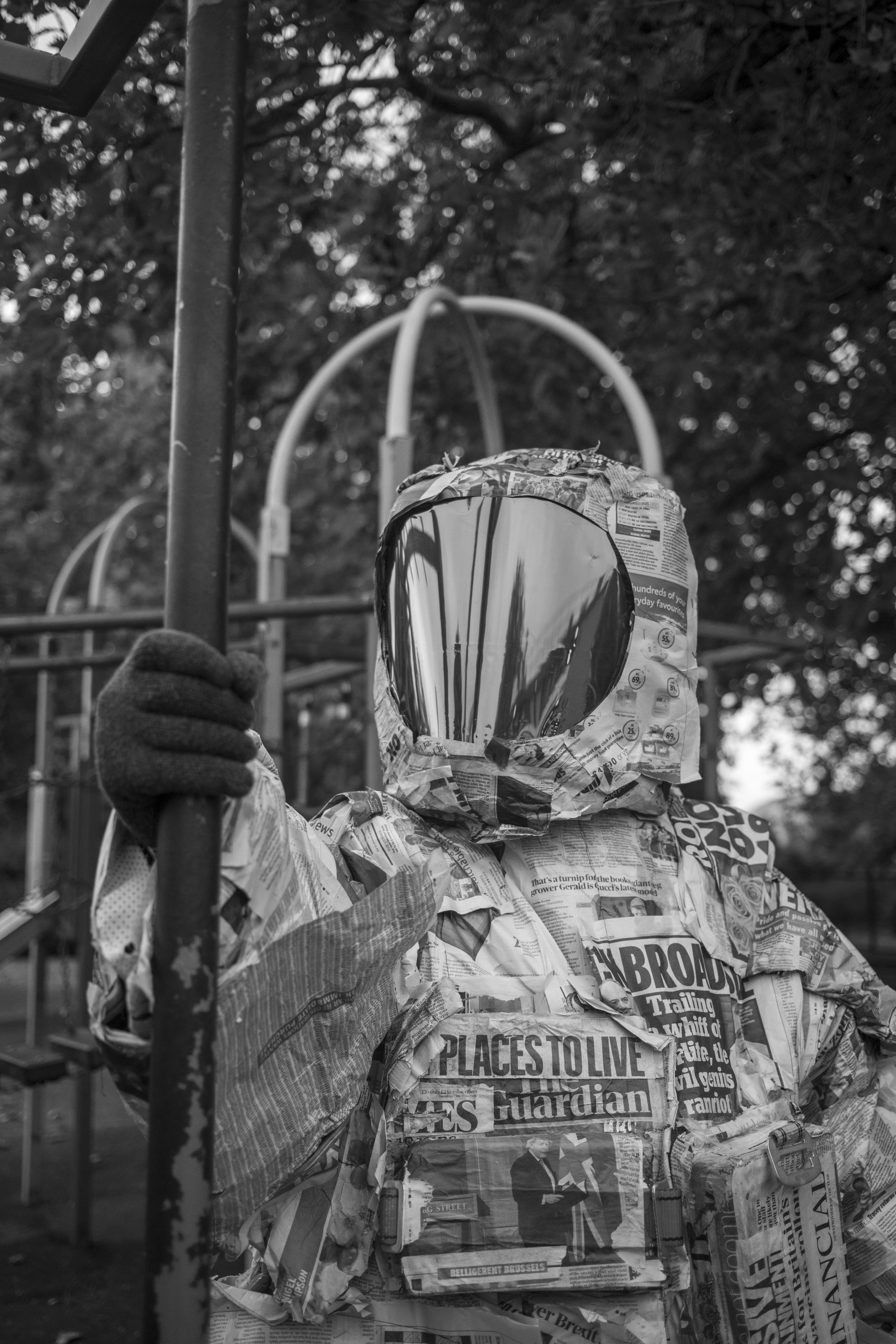 a black and white close-up photo of a person wearing a spacesuit made of newspaper, holding on to a lamppost against the backdrop of a park