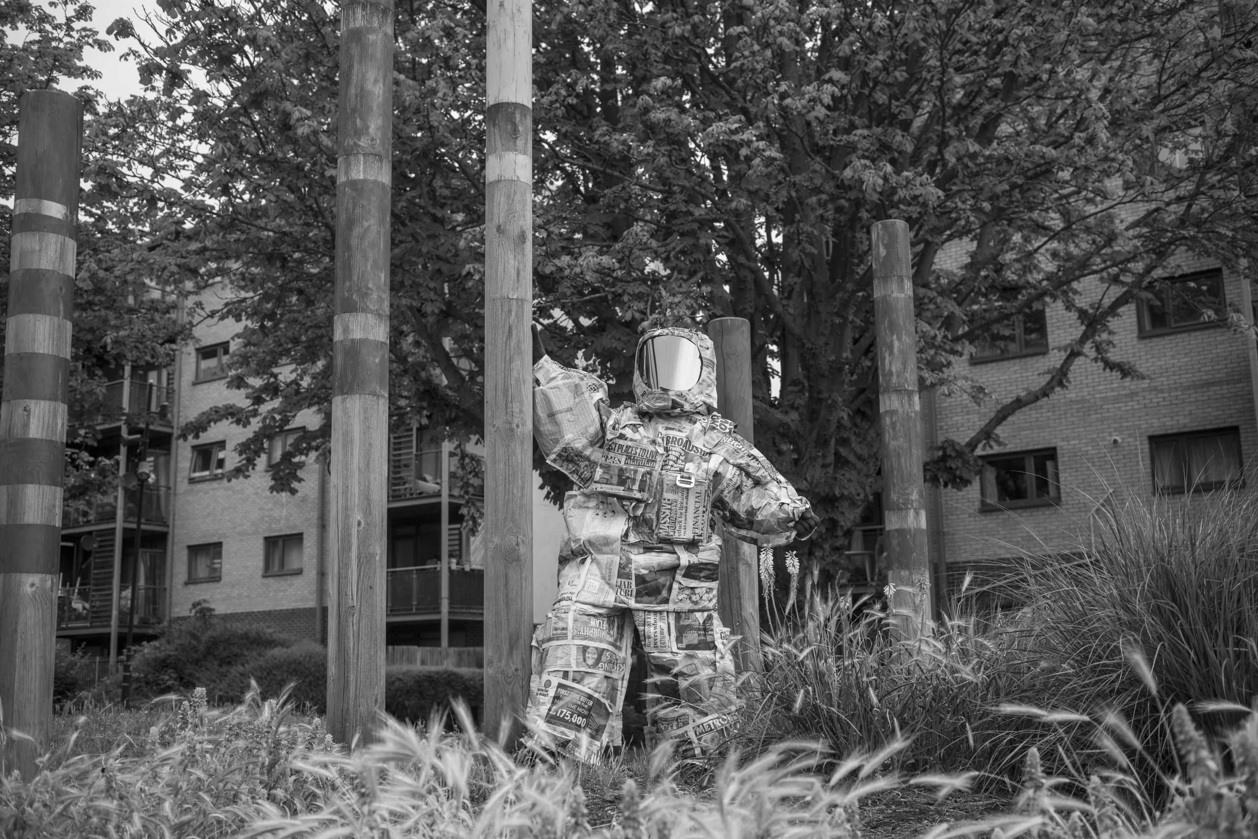 a black and white photo of some buildings, at the front we see someone dressed in what looks like a spacesuit that is made of newspapers, they are facing the camera and touching a wooden pole