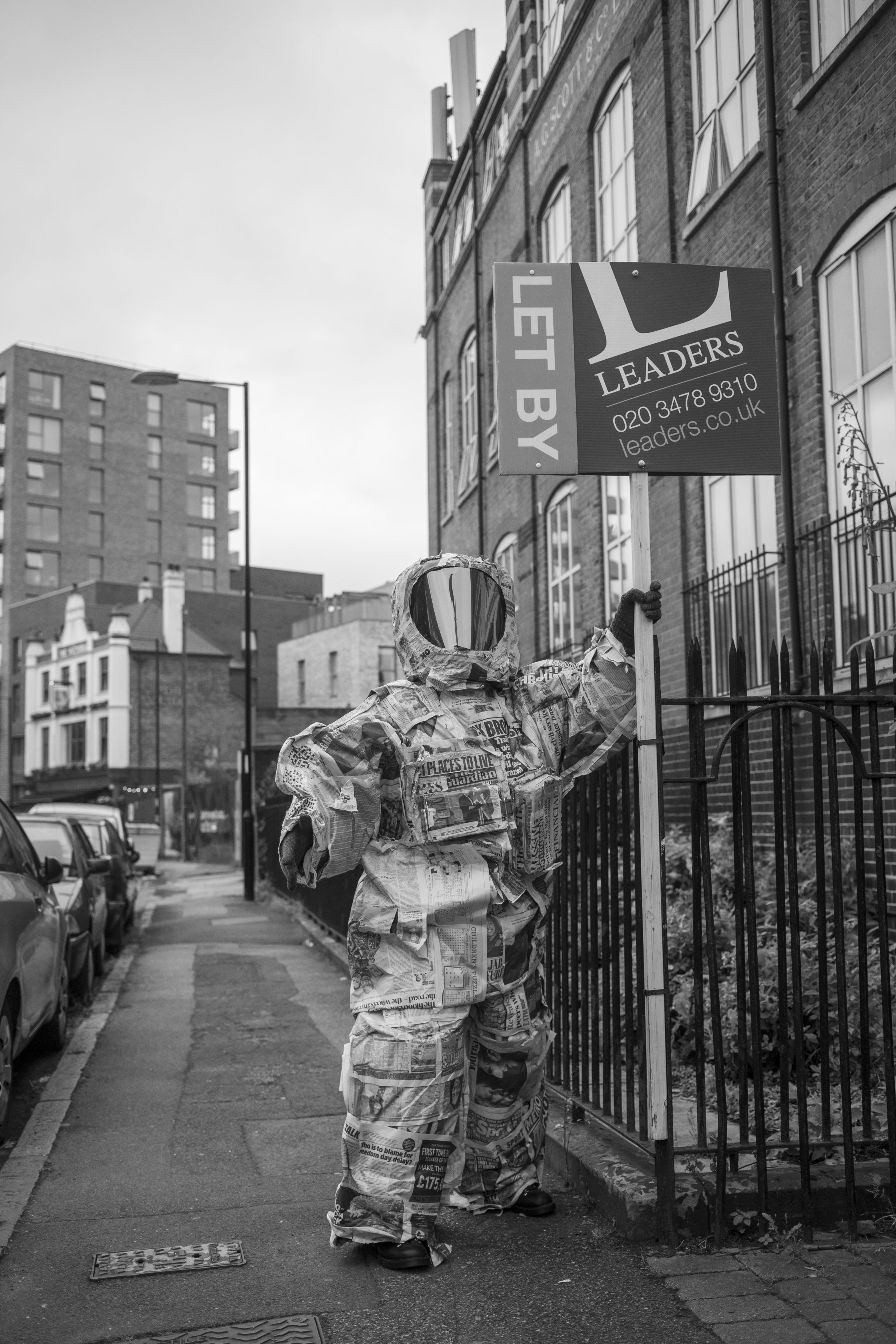 a black and white photo of a person wearing a spacesuit made of newspaper, they are holding a sign in front of a house that says 'Let By’ and ‘Leaders'