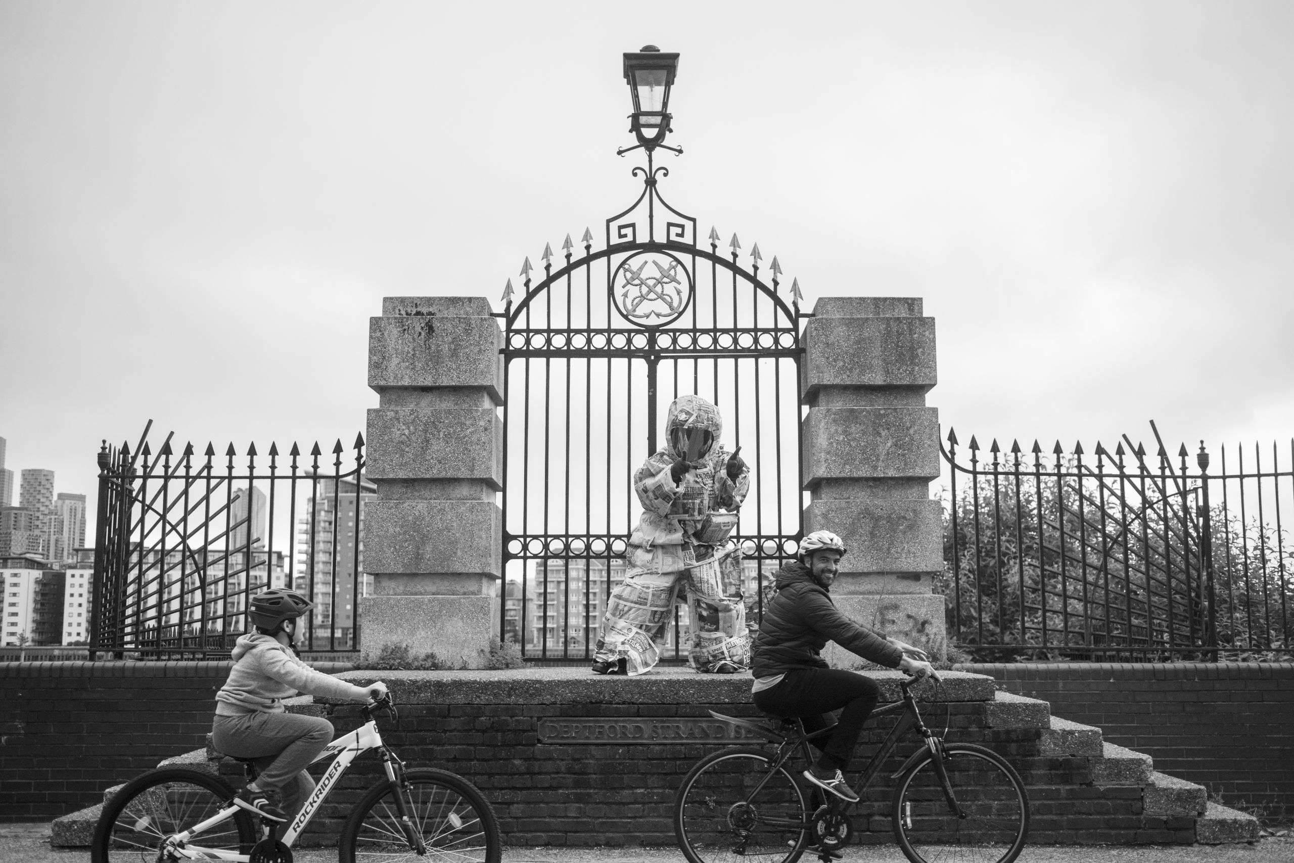a black and white photo of a person wearing a spacesuit made out of newspaper, standing in front of a gated platform, at the front, an adult cyclist and child cyclist cycle past with a grin on their face