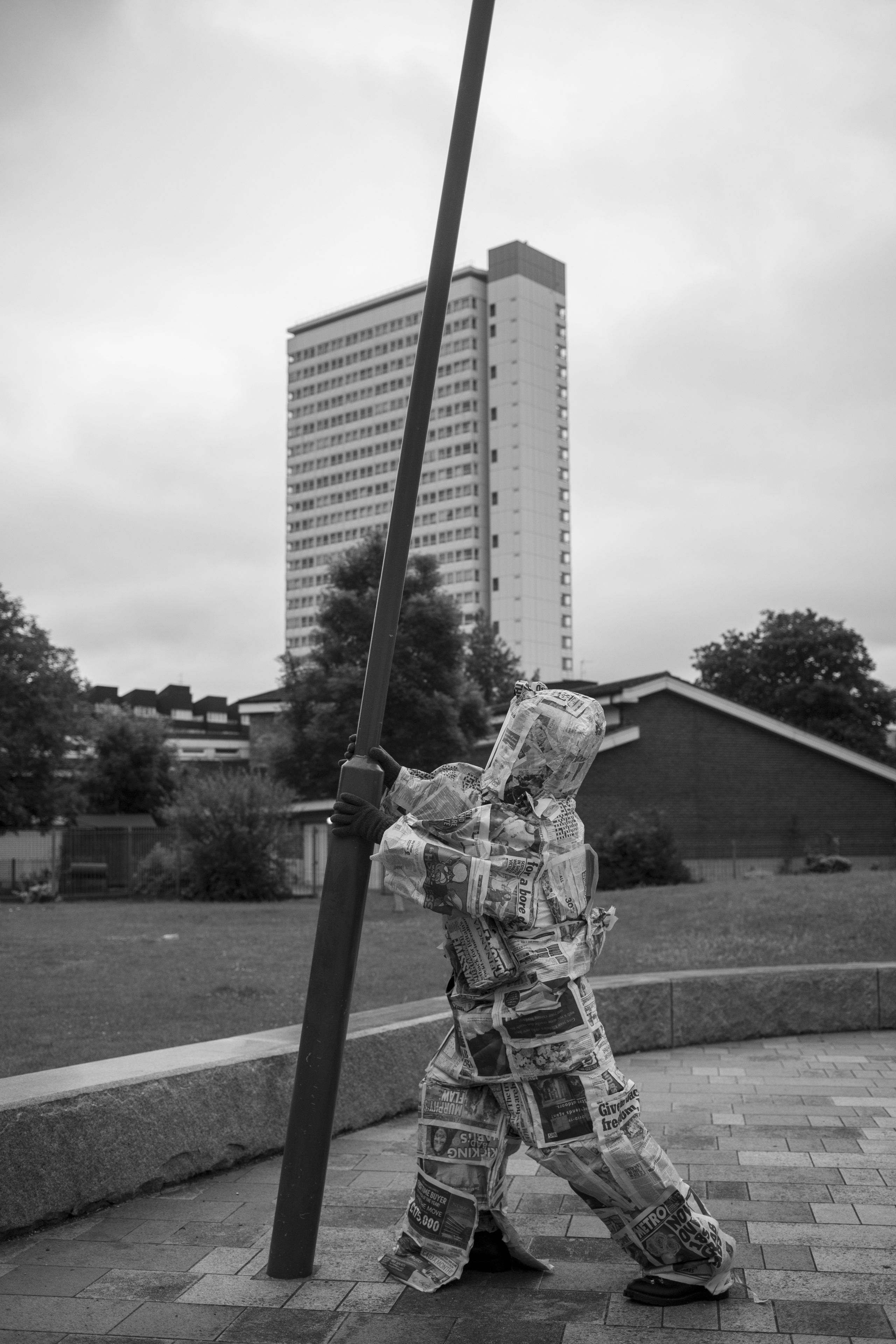 a black and white photo of someone wearing a spacesuit made of newspapers, holding on to a lamppost looking like they want to plant it in the grass, there is a building in the background