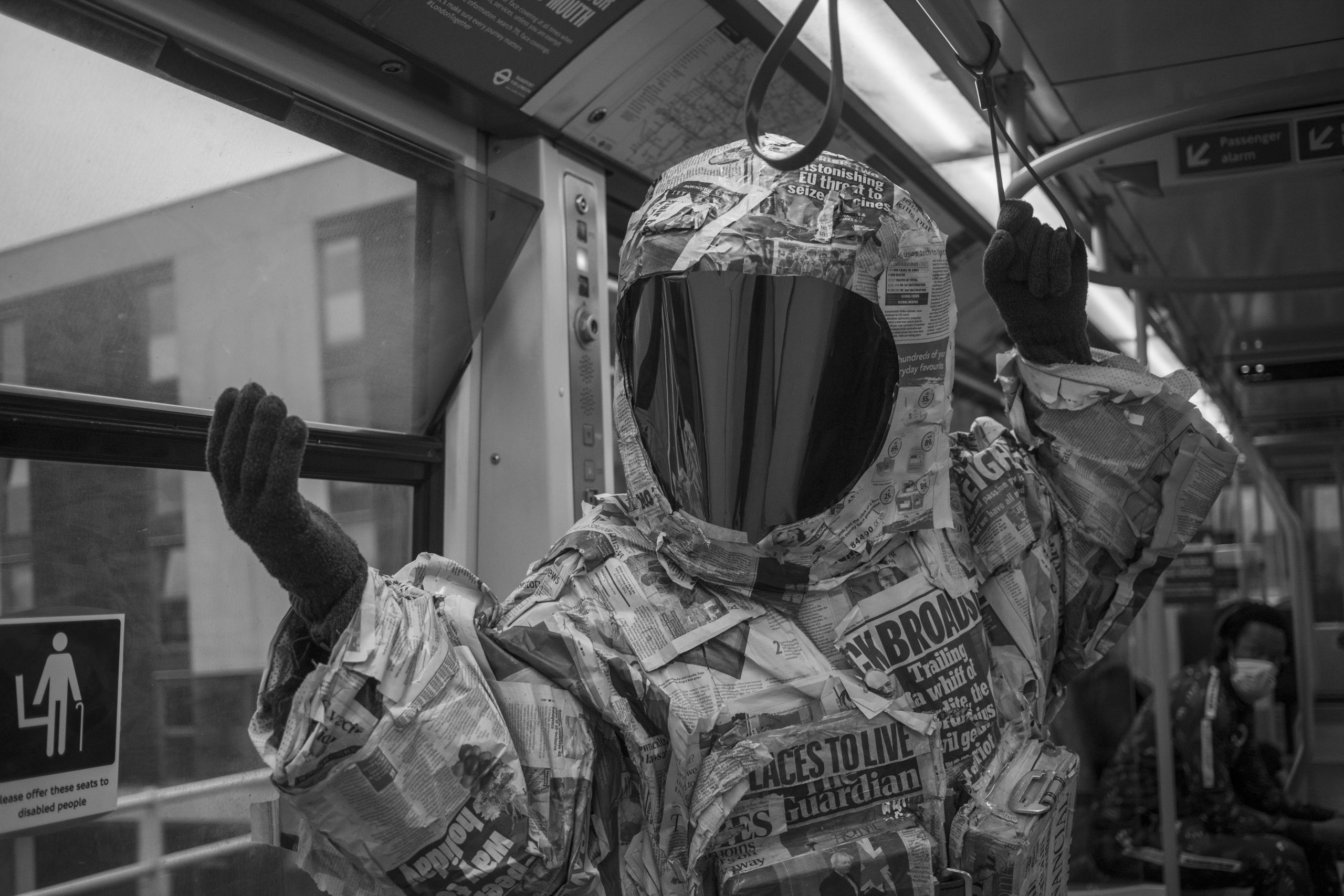 a black and white photo of a person wearing a spacesuit made of newspaper on an overground train, one hand is on the railing while the other hand is held out, extended in front of the train window