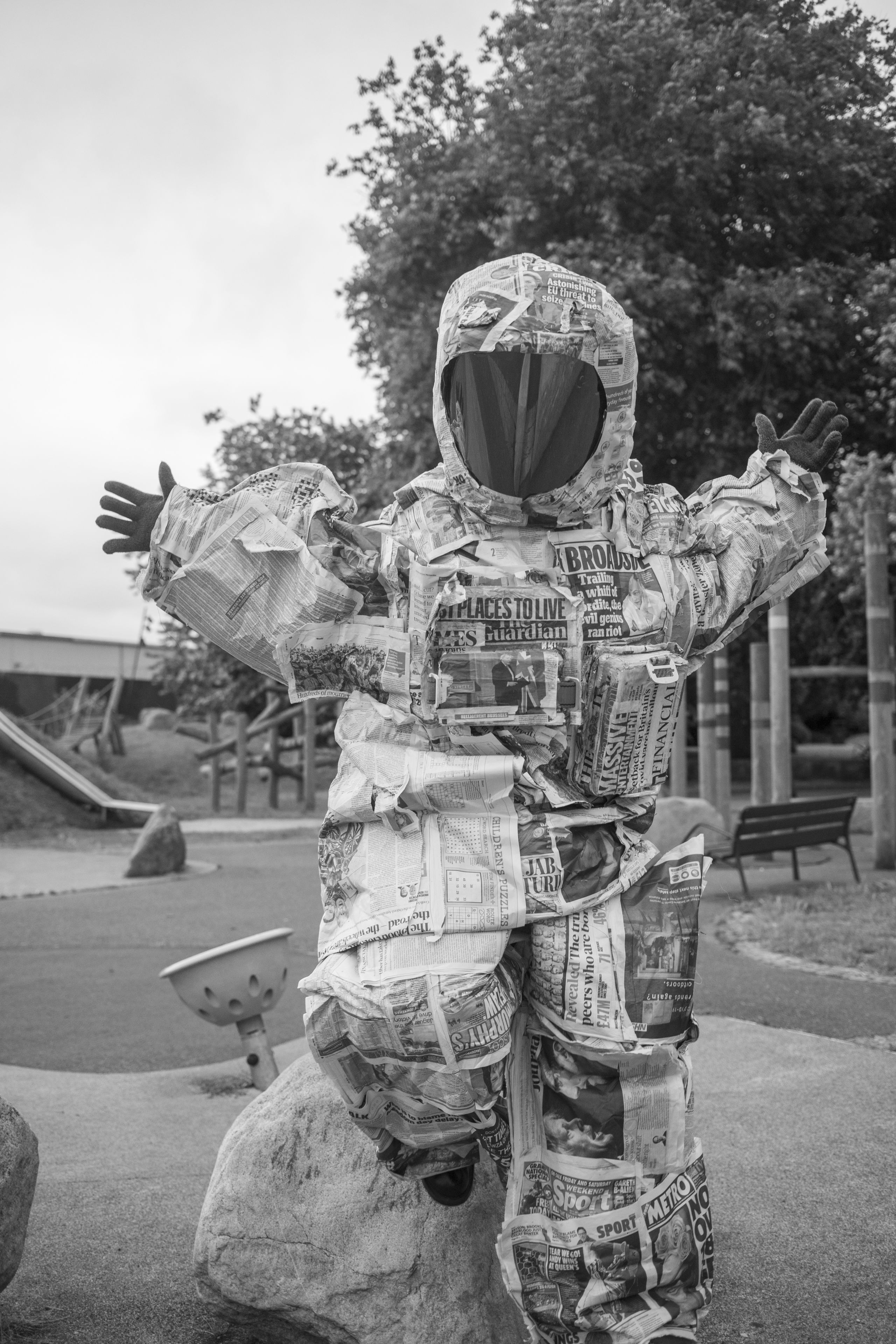 a black and white photo of a person wearing a spacesuit made of newspapers, shot in a playground, their arms extended out in the air