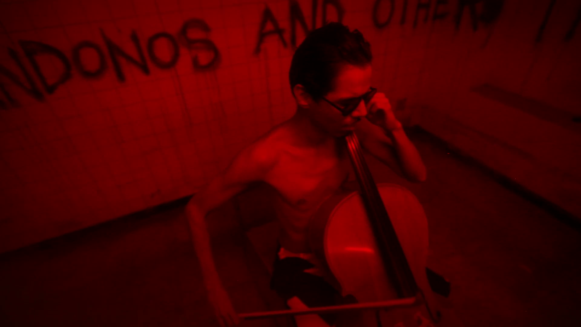 A topless man with black sunglasses plays the cello in a dirty bathroom, the photo has a deep red hue